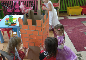 Cztery dziewczynki w bajkowych strojach przyklejają cegiełki do zamku z kartonu